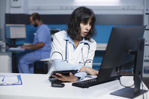 kvinna läkare kontroll patient uppgifter på dator medan gripande en läsplatta. i ett kontor, caucasian kvinna bär en labb täcka är använder sig av en digital enhet och skrivbordet pc till analysera medicinsk data. foto