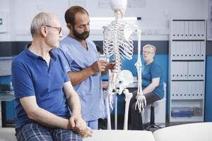 i fysioterapi klinik, ortoped förklarar spinal sladd på mänsklig skelett till äldre patient. läkare diskuterar anatomi och spinal ben med gammal man i förberedelse för osteopati diagnostisk. foto