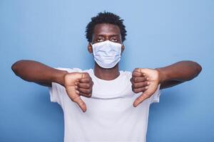 afrikansk amerikan vuxen som visar tummen ner tecken med både händer. svart man gestikulerar med hans fingrar medan ser på kamera och bär ansikte mask till skydda från coronavirus epidemi. foto