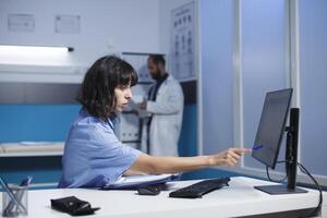 bild visar en sjuksköterska praktiker på de klinik skrivbord granskning och analyserar henne anteckningar. en caucasian kvinna i blå scrubs förbereder för patient medicinsk samråd förbi använder sig av en skrivbordet pc och urklipp. foto