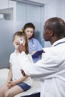 afrikansk amerikan läkare använder sig av termometer på liten flicka till kolla upp feber i medicinsk kontor. läkare åtgärder de temperatur till hitta en diagnos och ger behandling till de barn i en skåp. foto