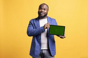 glad afrikansk amerikan man presenter på grön skärm läsplatta i landskap läge, studio bakgrund. optimistisk bipoc person innehav tom kopia Plats krom nyckel enhet Begagnade för visa upp annonser foto