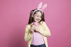 Lycklig upphetsad ung flicka Framställ med en fylld kanin i studio, bär kanin öron och innehav påsk dekoration i främre av de kamera. ung barn med pigtails varelse stolt av fluffig leksak. foto