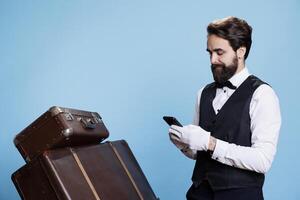 ung bellboy surfing webb på smartphone, verkande tycka om en professionell lyx hotell anställd på kamera. dörrvakt använder sig av internet webbplatser, granskning uppkopplad bokningar eller tjänster reservationer. foto