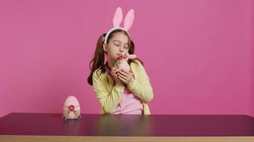 leende litet barn med kanin öron som visar henne handgjort påsk ornament, presenter en handgjord dekorerad ägg och kanin. ung ljuv skol innehav festlig dekorationer. kamera b. foto