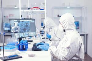forskning forskare skriver information erhållna arbetssätt i modern laboratorium. medicinsk ingenjör använder sig av dator under global pandemi med coronavirus klädd i overall. foto
