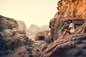 caucasian ung kvinna turist sitta och leende medan ser till skön petra gammal stad sniderier. känd besök jordan attraktion destination. petra arkeologisk webbplats panorama foto