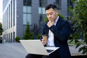 allvarlig och omtänksam ung asiatisk affärsman Sammanträde på en bänk nära ett kontor byggnad bär hörlurar, innehav en kopp av kaffe och ser på en bärbar dator övervaka. foto