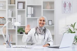 en glad kvinna läkare i hijab vågor till de kamera medan Sammanträde på en skrivbord i en ljus, välorganiserad kontor. teknologi och sjukvård professionalism kombinerad. foto
