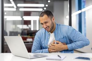 ett indisk man i en blå skjorta visas bedrövad eller i obehag på hans kontor, gripande hans bröst medan arbetssätt. foto