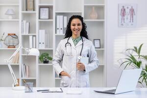 porträtt av en leende latin amerikan kvinna läkare stående i en sjukhus kontor i en vit täcka och innehav en läsplatta, ser och leende på de kamera. foto
