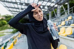 ett utmattad kvinna idrottare i hijab avtorkning svettas från henne panna med en flaska av vatten på en stadion. foto