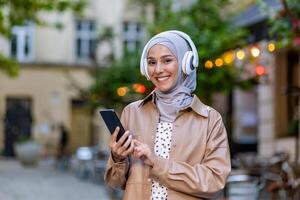 charmig muslim kvinna bär hörlurar över hijab och använder sig av mobil telefon under promenad i stad. positiv eleganta lady njuter lyssnande till favorit musik genom elektronisk trådlös enheter. foto