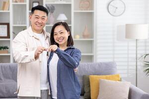 ung Lycklig asiatisk par man och kvinna leende och ser på kamera tillsammans, innehav nycklar till deras ny lägenhet hus, porträtt av Lycklig Hem ägare. foto