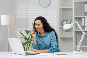 en fokuserade kvinna i en blå skjorta använder sig av en headsetet medan arbetssätt på en bärbar dator i en ljus kontor miljö, engagerande i en företag diskussion. foto