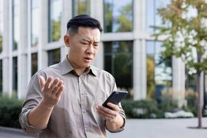 orolig senior asiatisk manlig kontor arbetstagare affärsman stående utanför och använder sig av mobil telefon. besvikelse utseende på de skärm. foto