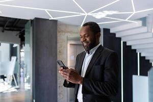 framgångsrik afrikansk amerikan affärsman inuti kontor stående förbi fönster, chef använder sig av telefon, man skriver meddelande leende och läsning uppkopplad Nyheter, skriver meddelande i social nätverk app. foto
