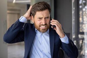 närbild Foto av en ung affärsman man i en kostym stående i ett kontor Centrum, oroligt talande på de telefon och innehav hans huvud med hans hand i frustration, lösning finansiell problem.