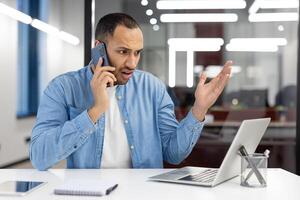 en man är talande på hans cell telefon medan Sammanträde på en skrivbord med en bärbar dator och en anteckningsbok. han är upprörd eller arg, eventuellt på grund av till en arbetsrelaterad problem foto