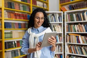 leende ung kvinna i glasögon använder sig av en digital läsplatta bland bokhyllor i en bibliotek, representerar modern inlärning och teknologi i utbildning. foto