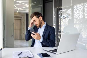 chockade ung man affärsman Sammanträde i de kontor på en stol och innehav hans huvud med hans hand, innehav de telefon, upprörd förbi de Nyheter han mottagen. foto