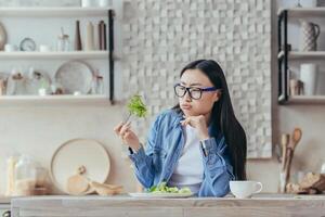porträtt av en ung skön asiatisk kvinna Sammanträde i de kök på de tabell, innehav en färsk grön sallad i henne gaffel, suckar, ser på den olyckligt. trött av bantning, hatar vegetarianism. foto