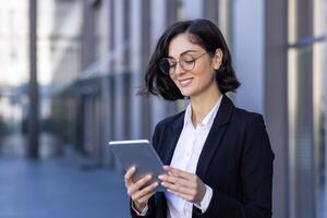 en leende ung affärskvinna är stående nära ett kontor byggnad och använder sig av en läsplatta. samtal på en ringa upp, texter, närbild foton. foto