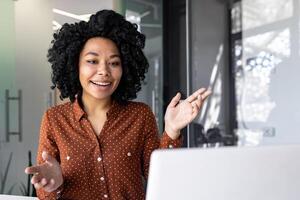 glad framgångsrik affärskvinna talande avlägset använder sig av bärbar dator för ringa upp, kvinna anställd leende och gestikulerar ser på dator skärm, arbetssätt inuti kontor på arbetsplats. foto