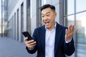 Lycklig asiatisk affärsman stående utanför i en kostym och överraskad och Lycklig ser på de skärm av hans mobil telefon. foto