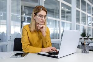 porträtt av en allvarlig tänkande affärskvinna inuti en ljus kontor på arbete med en bärbar dator, en kvinna i en gul skjorta och glasögon Arbetar eftertänksamt medan Sammanträde på en arbetsplats foto