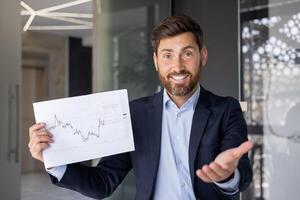 leende, professionell affärsman innehav en finansiell Graf, gestikulerar ett inbjudande hand, indikerar tillväxt i en modern kontor miljö. foto