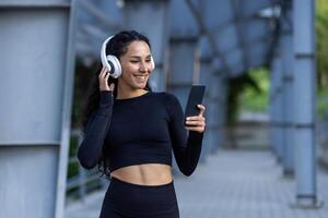 latinamerikan sports med hörlurar lyssnande till uppkopplad musik och audio podcasts med böcker medan joggning och håller på med aktiva övningar, kvinna använder sig av app på telefon, i träningsoverall. foto