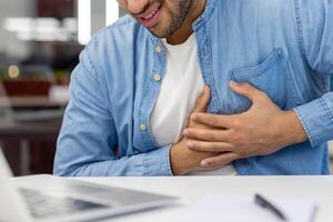 en närbild Foto i de arbetssätt kontor av en ung mannens händer innehav hans bröst, känsla en stark smärta i hans hjärta.
