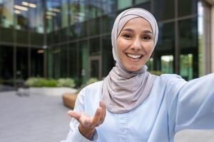 en glad kvinna bär en hijab är tar en selfie med en smartphone i ett urban miljö, strålande positivitet och förtroende. foto