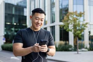 en ung man, ett asiatisk sportsman, är stående på en stad gata i hörlurar, innehav en telefon i hans händer, framställning en ringa upp. rattar meddelanden, chattar. foto