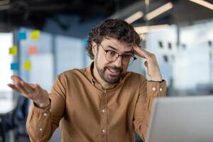 förvirrad professionell manlig bär glasögon, uttrycker förvirring medan använder sig av en bärbar dator i en ljus samtida kontor Plats. foto