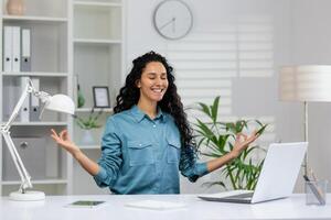 leende affärskvinna med ögon stängd praktiserande yoga meditation på henne skrivbord i en ljus kontor miljö till minska påfrestning. foto