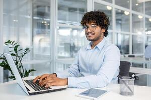 en ung professionell man med lockigt hår, bär glasögon och en ljus blå skjorta, är arbetssätt på hans bärbar dator i en ljus, modern kontor miljö. han utseende fokuserade och innehåll. foto