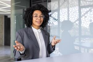 ung afrikansk amerikan företag kvinna håller på med Träning, uppkopplad möte Sammanträde i kontor på tabell, talande och leende på kamera. foto