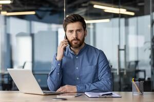 porträtt av en allvarlig ung affärsman Sammanträde i de kontor på de tabell i främre av de kamera och talande på de telefon. foto