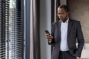 fokuserade mogna afrikansk amerikan affärsman i en kostym använder sig av hans smartphone i en modern kontor miljö. foto