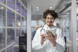en läkare i en vit medicinsk täcka och stetoskop innehar en läsplatta för en medicinsk rådfråga i hans kontor. leende man ser på kamera foto