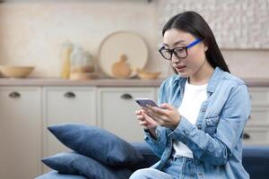 ung skön kvinna på Hem Sammanträde i de kök leende och Lycklig, asiatisk kvinna använder sig av smartphone skriver meddelanden och läsning Nyheter uppkopplad foto