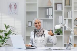 en glad kvinna läkare bär en hijab sitter på henne skrivbord i en klinik, ger en tummen upp medan arbetssätt på henne bärbar dator, omgiven förbi medicinsk böcker och växter. foto