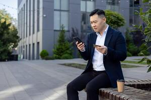 upprörd ung asiatisk manlig kontor arbetstagare Sammanträde på en bänk under en arbete ha sönder, innehav en kreditera kort och ser på de telefon i frustration. foto