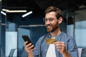 glad och leende skäggig affärsman håller på med uppkopplad handla och Bank pengar överföra, frilansare innehav Bank kreditera kort och smartphone, man Sammanträde på skrivbord inuti kontor. foto