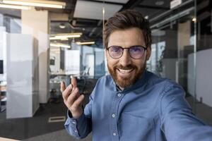 närbild Foto av en ung leende man i de kontor talande på en ring upp till de kamera telefon, förklara förbi gestikulerar med hans hand.