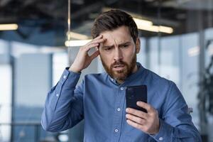 upprörd och allvarlig ung manlig affärsman stående i kontor och ser chockade på telefon skärm, innehav hand till huvud. foto