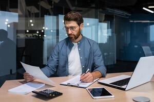 allvarlig och fokuserade finansiär revisor på papper arbete inuti kontor, mogna man använder sig av kalkylator och bärbar dator för beräknande rapporter och sammanfatta konton, affärsman på arbete i tillfällig kläder. foto