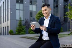 asiatisk leende ung man Sammanträde utanför ett kontor Centrum bär en headsetet och bläddring på en läsplatta. foto